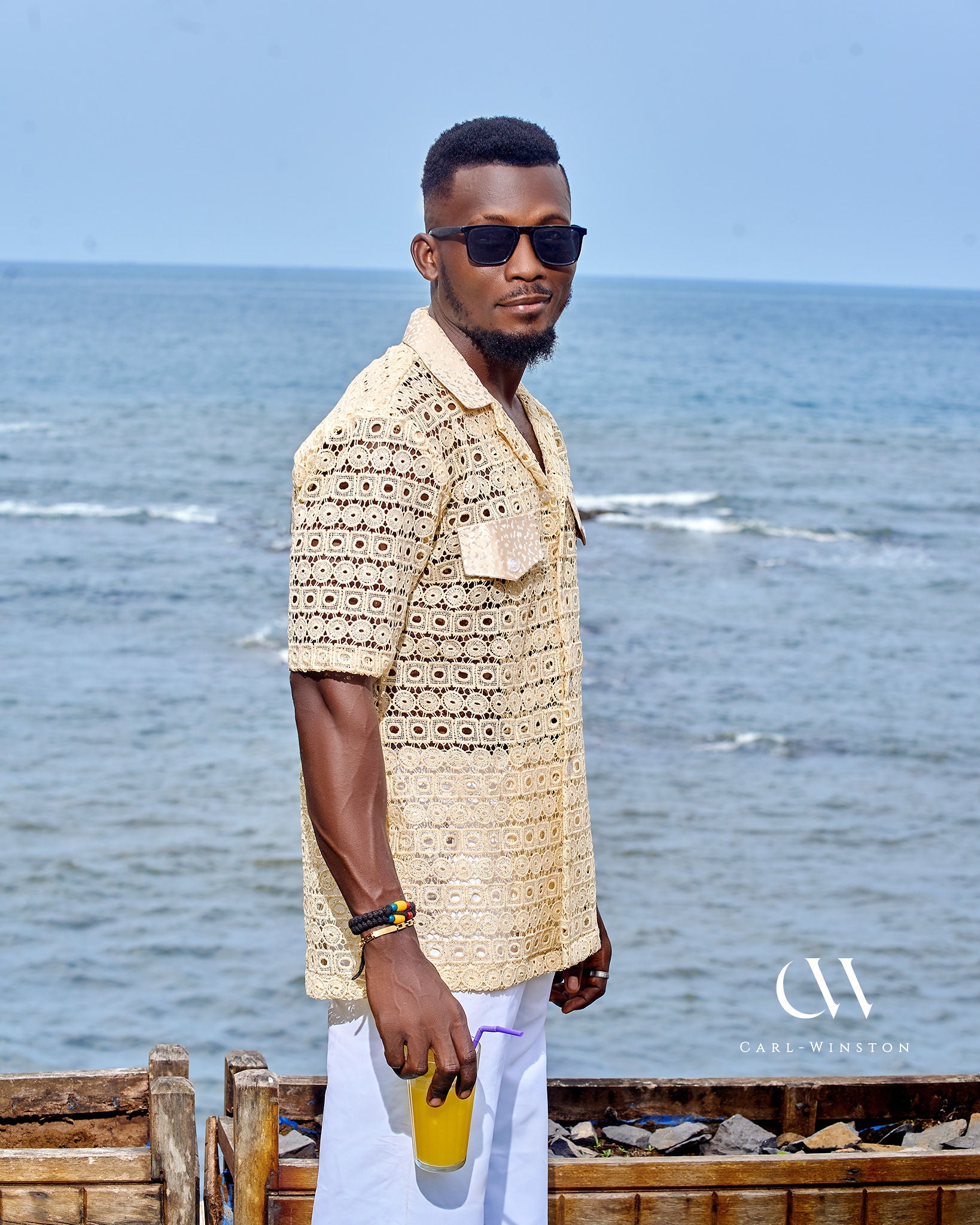 Man holding a tropical drink while modeling Carl-Winston’s lightweight lace shirt against a seaside backdrop.