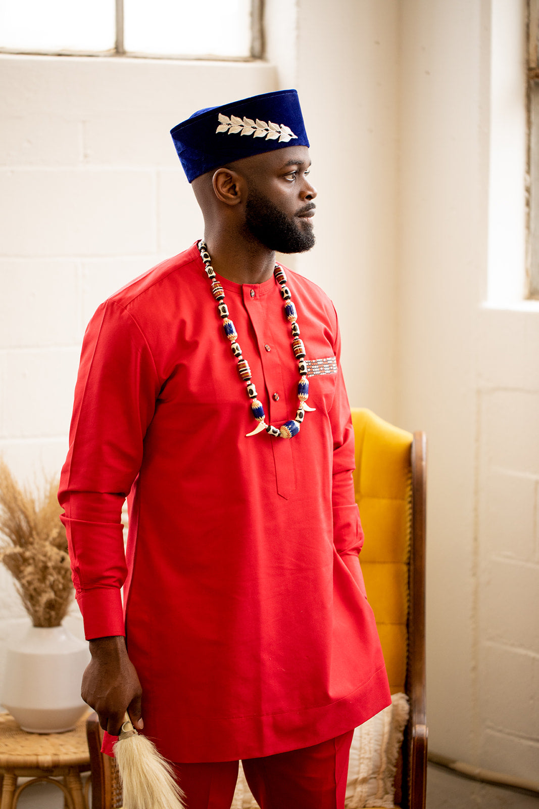 Carl-Winston traditional men's attire featuring a bold red outfit with a striking blue velvet hat adorned with silver leaf details. This look is completed with an ornate beaded necklace and an animal tail staff, embodying elegance and cultural heritage, perfect for special occasions.