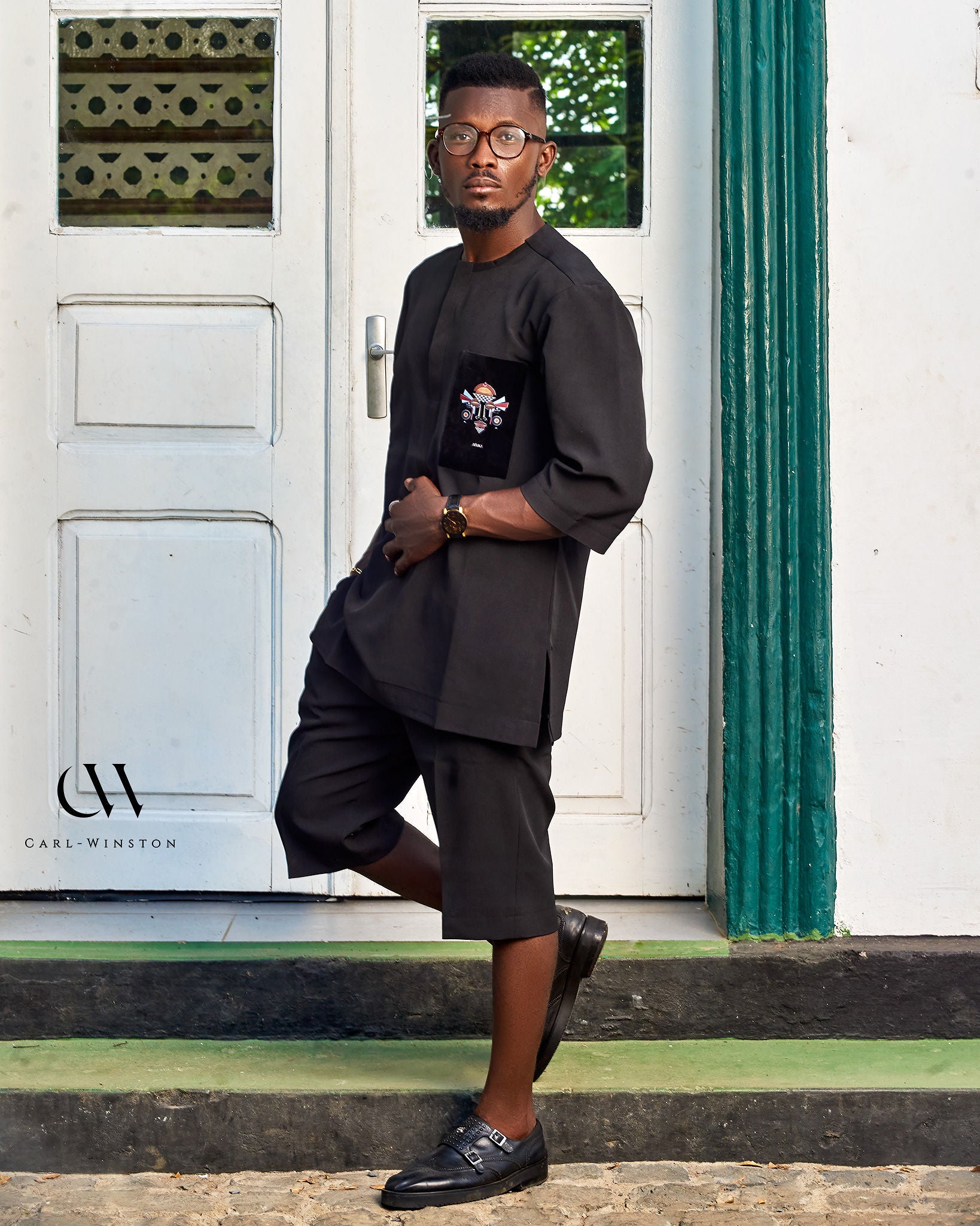 Side profile of a male model in a black shirt and tailored long shorts, standing in front of a white and green colonial-style building