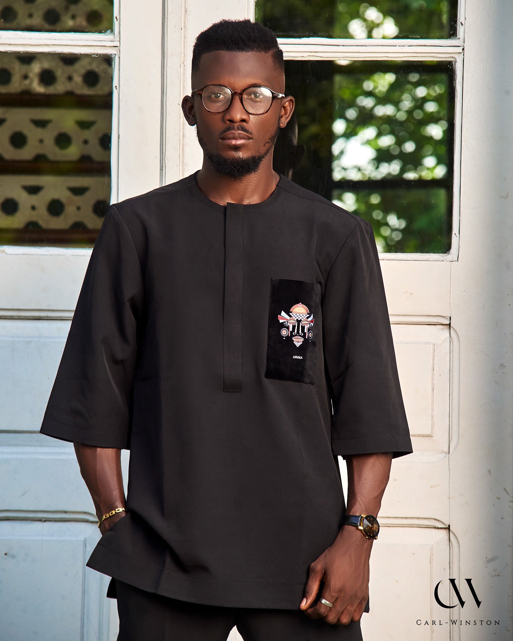 Front view of a male model wearing a black cotton shirt with 3/4 sleeves, featuring an embroidered velvet chest patch with a stylized African mask and the word “AFRIKA”