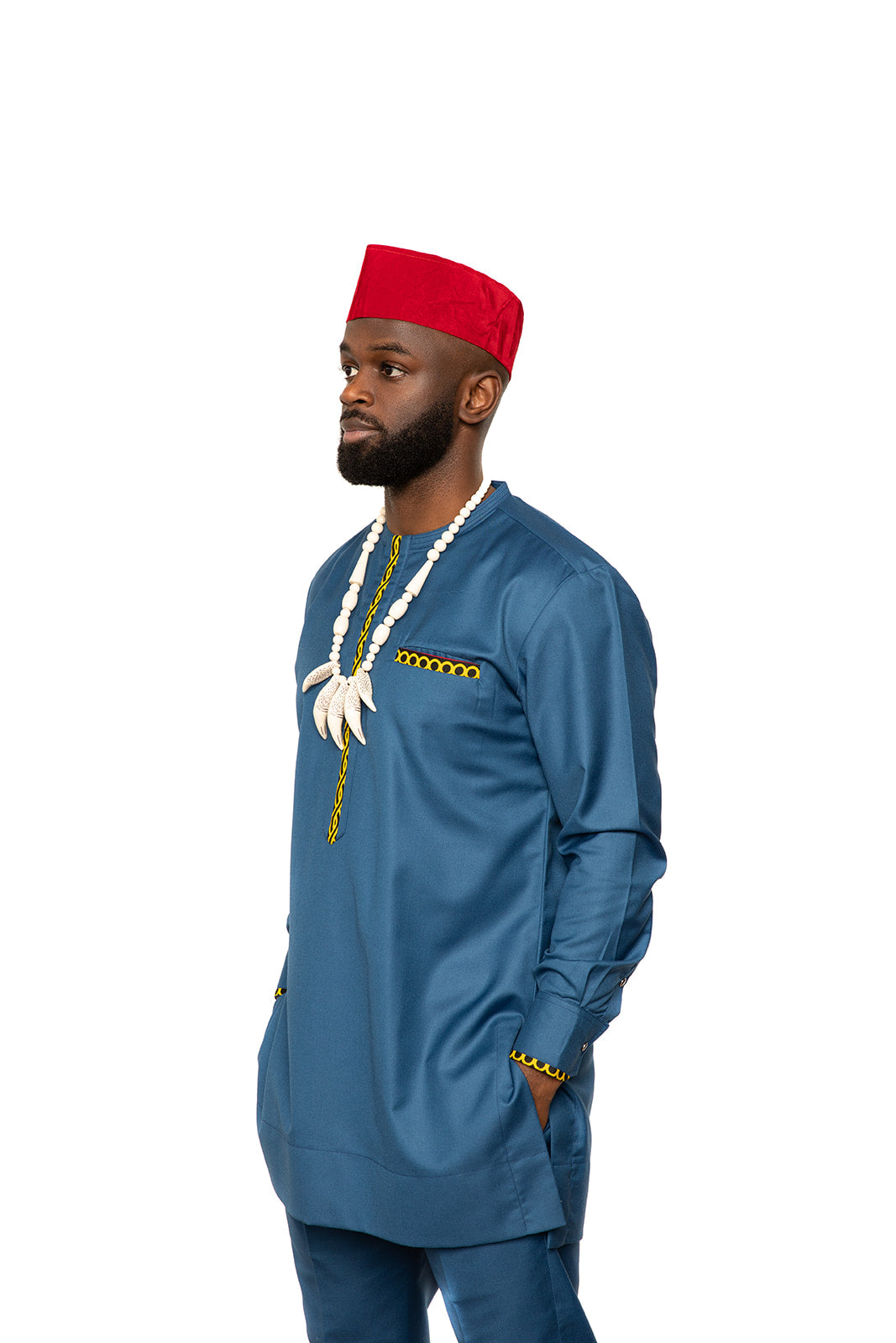 Carl-Winston traditional men's attire in a striking blue outfit paired with a red velvet cap and an ivory bead necklace adorned with tusk-shaped pendants. This ensemble is completed with yellow accents on the pocket, neckline, and cuffs, adding a unique touch to the elegant and culturally inspired design. Perfect for special occasions and formal events.