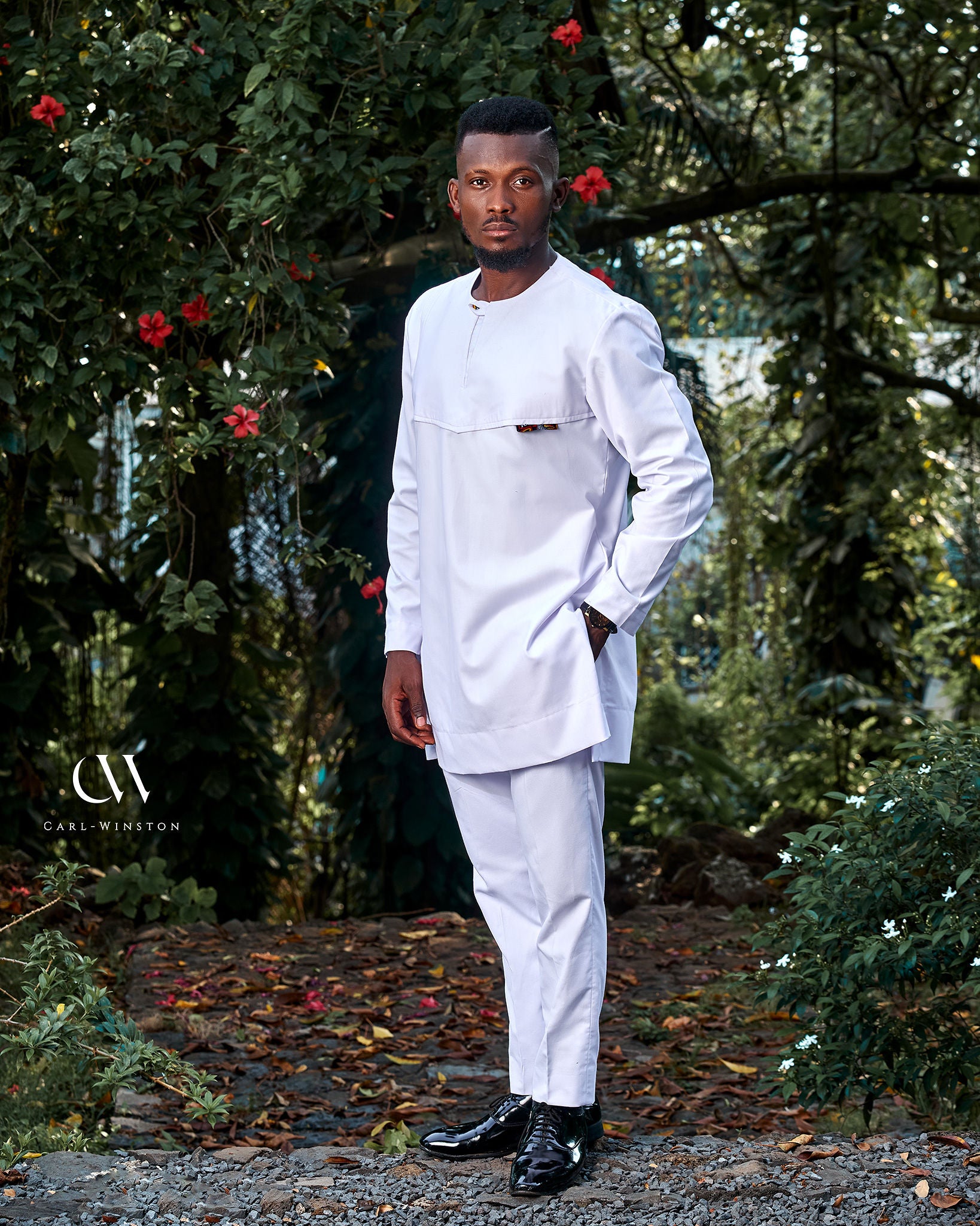 A confident African man standing in a vibrant, sunlit garden wearing a crisp white traditional Cameroonian outfit with colorful geometric Atoghu embroidery on the chest. Lush tropical greenery and blooming red hibiscus flowers surround him. His look is poised and elegant, symbolizing celebration and cultural pride during Palm Sunday or Easter. Professional fashion photography style.