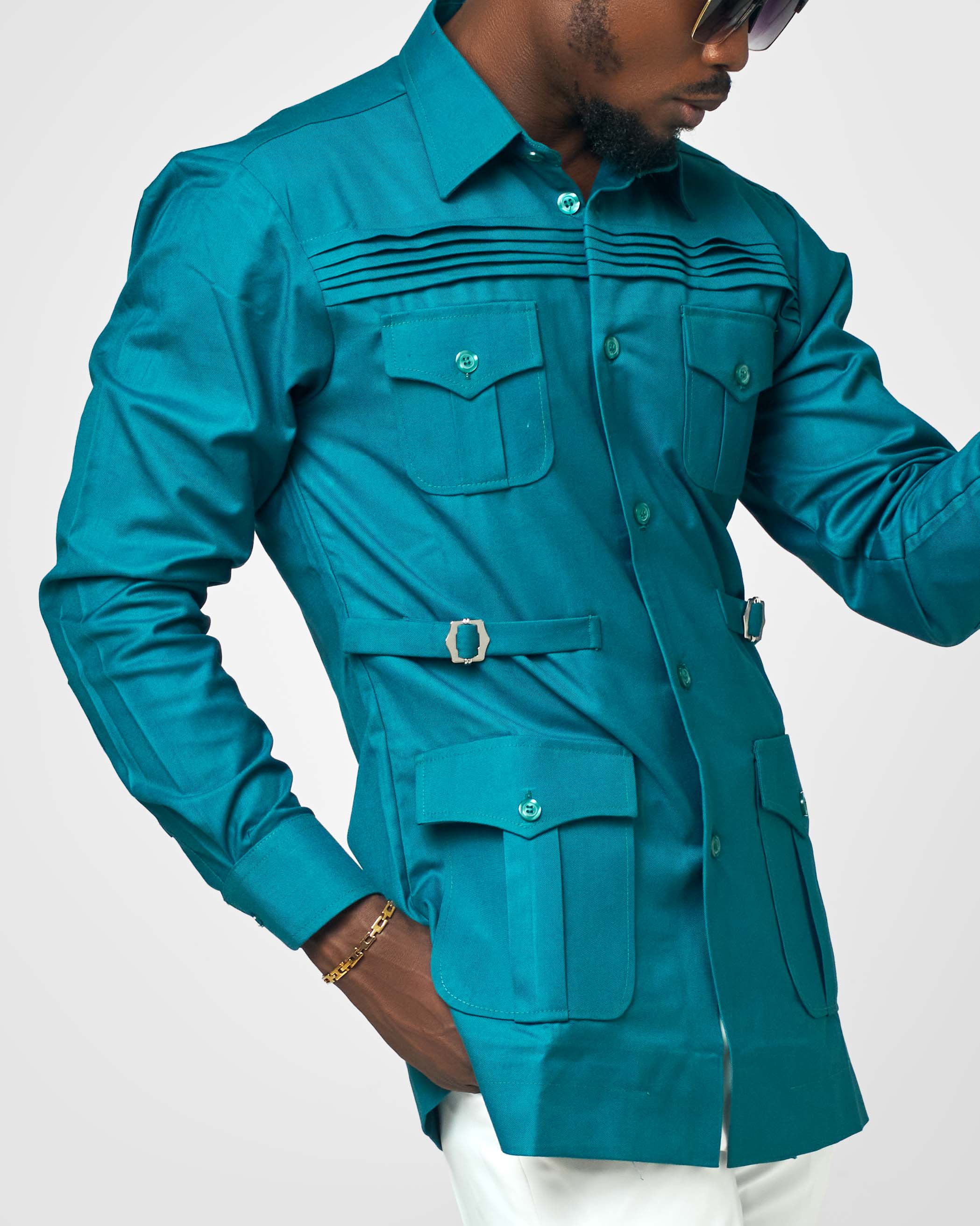 A modern teal men's utility shirt with front pockets, side belt accents, and pleated detailing across the chest, styled by Carl-Winston.