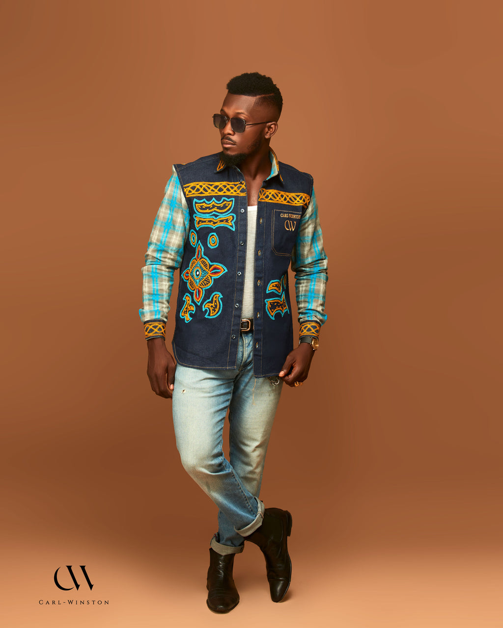 Man wearing a patterned vest over a plaid shirt with jeans and boots on a brown background