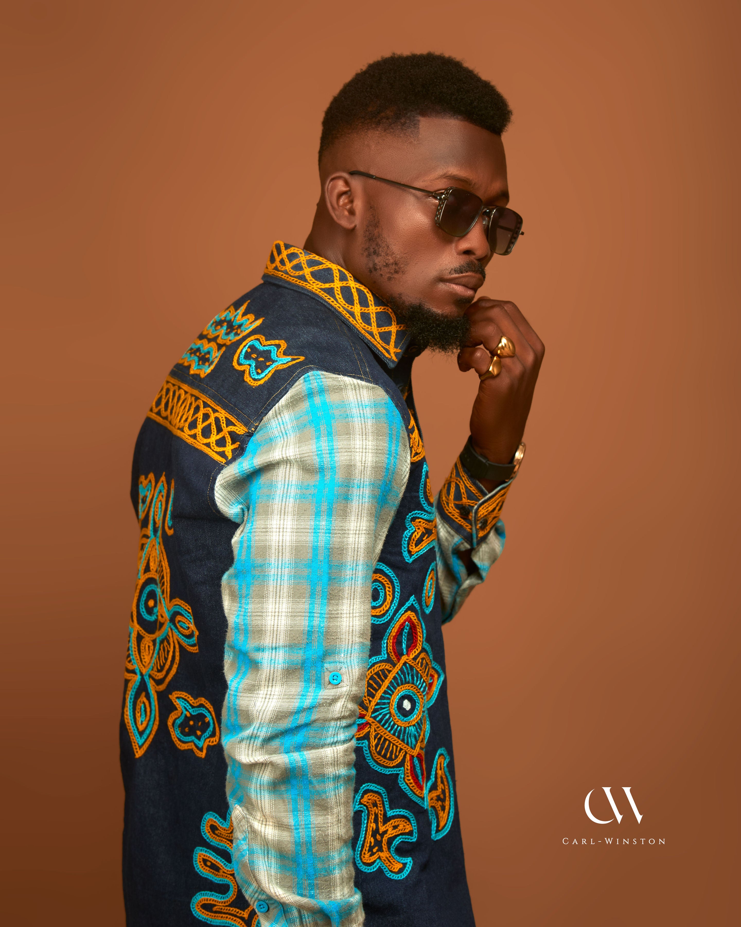 Man wearing an ornate jacket with a plaid shirt underneath against a brown background