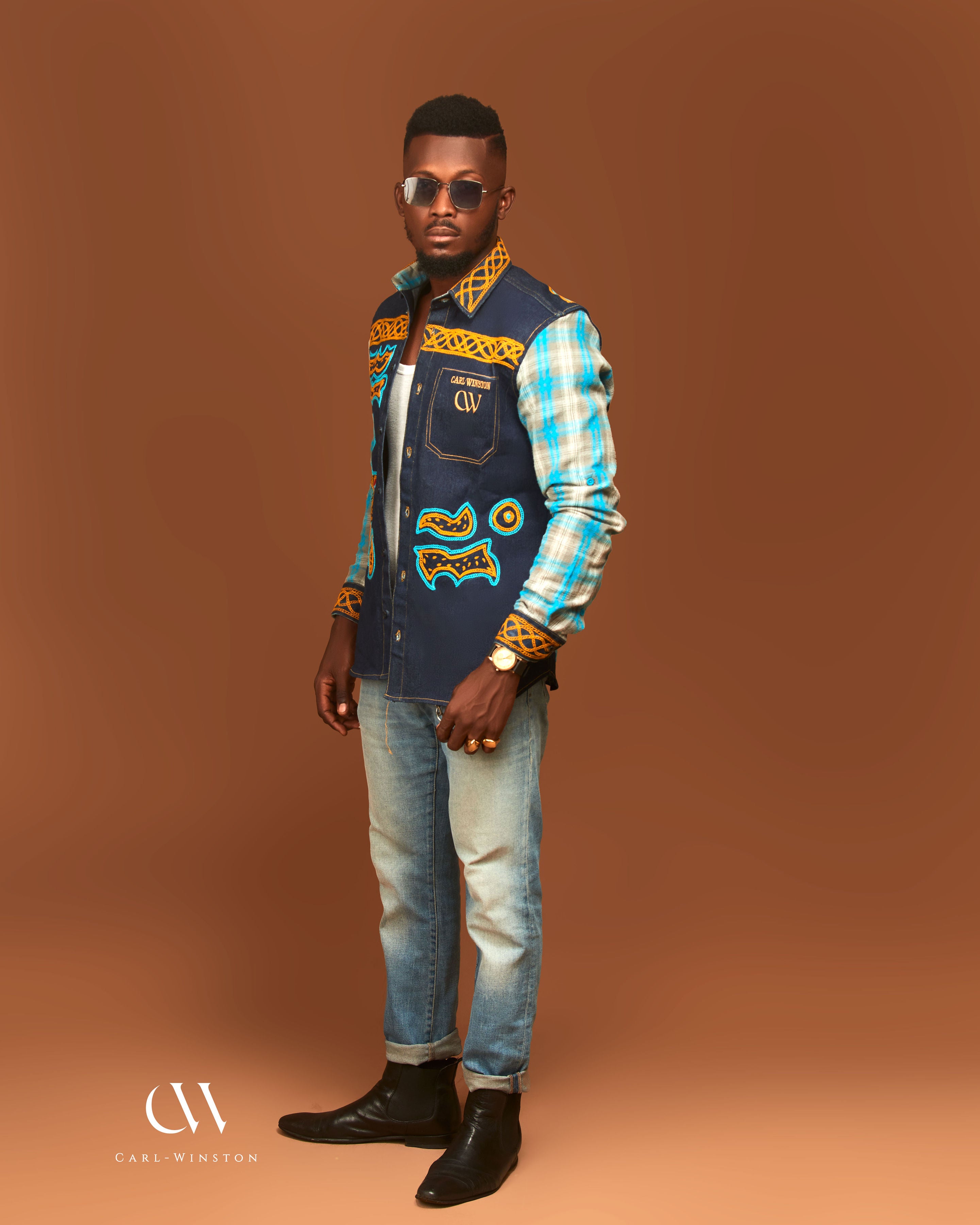 Man wearing a colorful jacket with patterns, plaid shirt, and jeans on a brown background