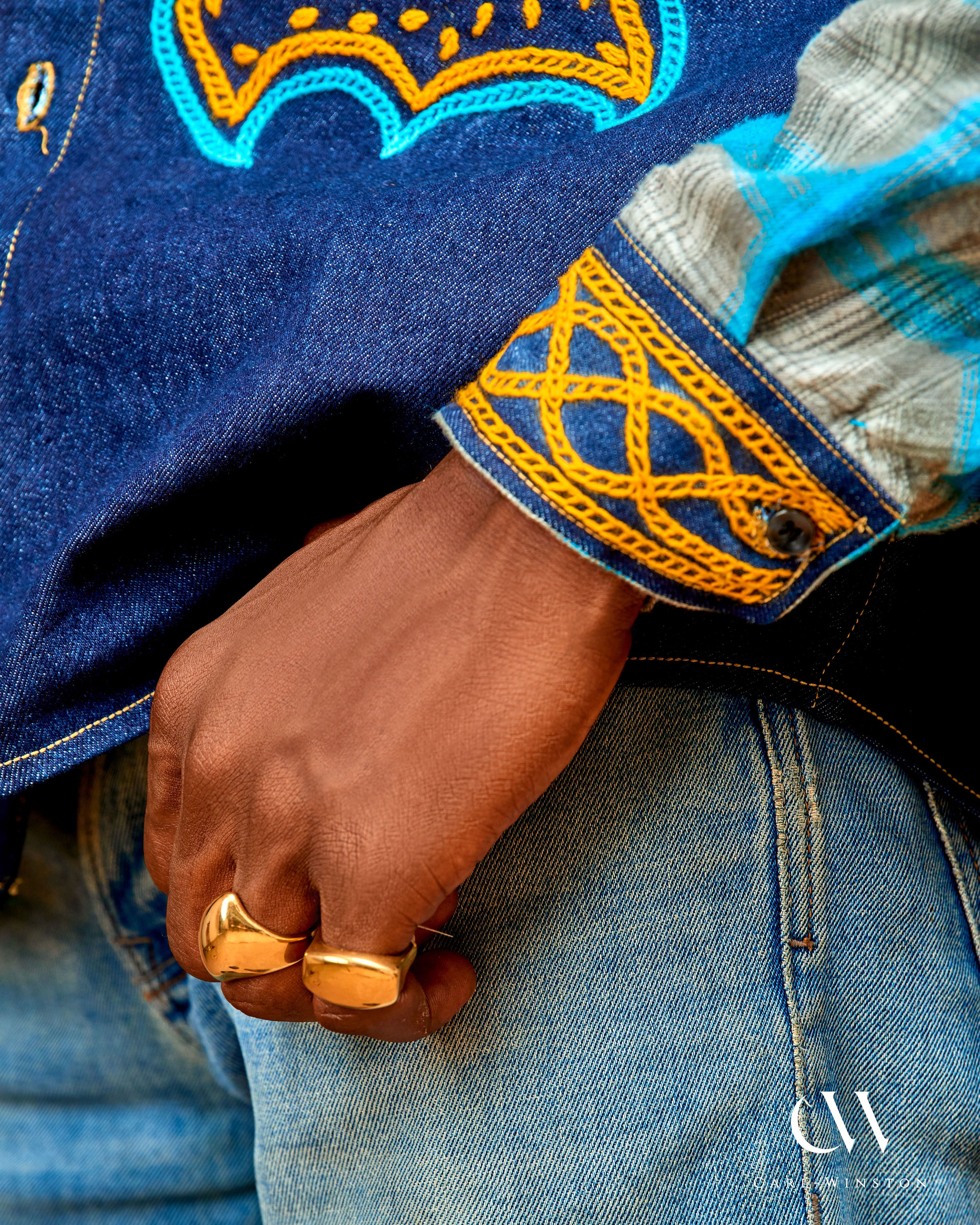Close-up of a hand wearing a gold ring on blue denim fabric with intricate yellow embroidery.