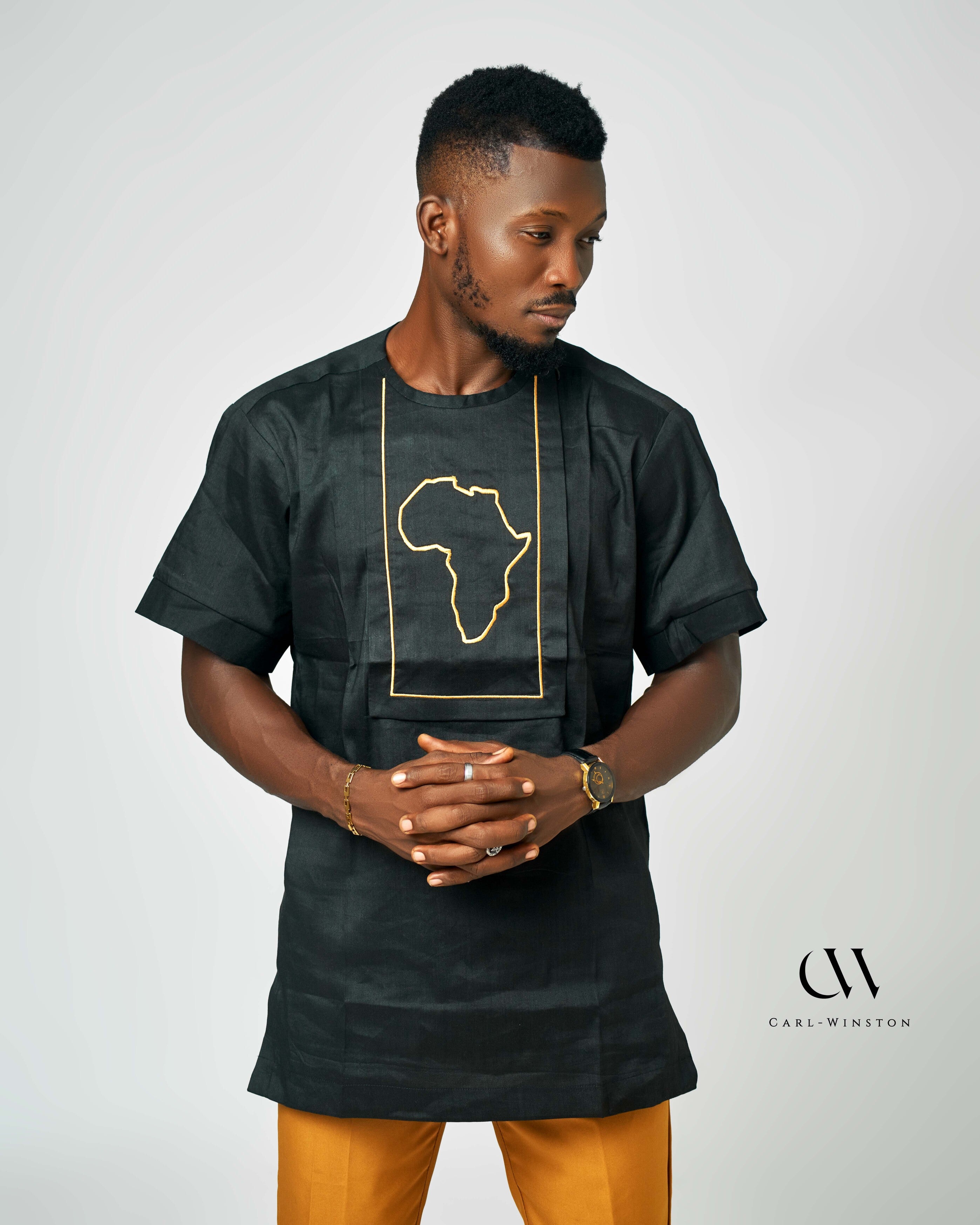 Man wearing a Black short-sleeve linen shirt with an embroidered golden outline of the African continent, paired with mustard-yellow pants, showcasing bold and cultural fashion.
