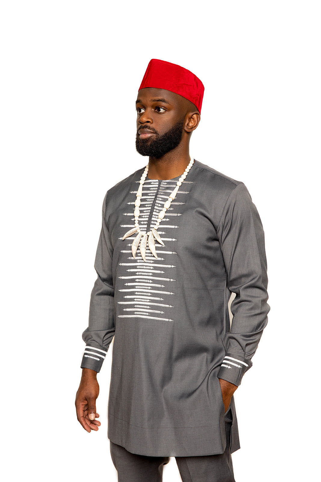 A Black man with a beard wearing a traditional African outfit stands confidently with his hand in his pocket. He is dressed in a dark gray long-sleeve tunic adorned with white embroidered patterns down the front and on the cuffs. He wears a red traditional hat and a bold beaded necklace featuring claw-like pendants, adding a touch of cultural significance to the ensemble. His expression is calm and poised as he looks off into the distance.