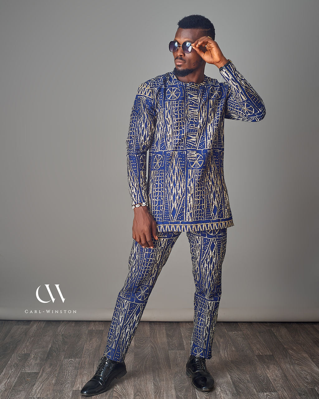 Man wearing a blue and white patterned outfit with sunglasses, standing against a gray background.