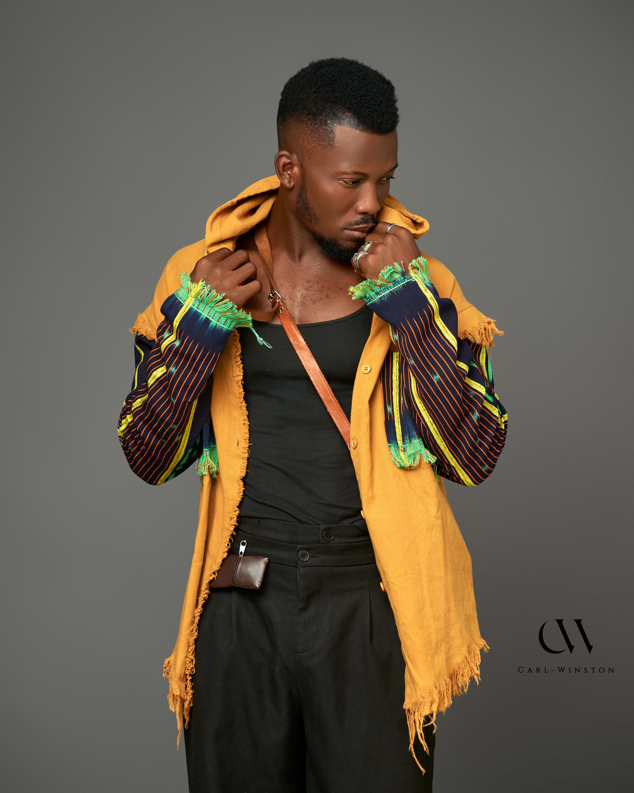 A striking Heritage Threads Hooded Jacket featuring distressed mustard-colored cotton with bold, woven African fabric sleeves, modeled by a stylish man in a black tank top.