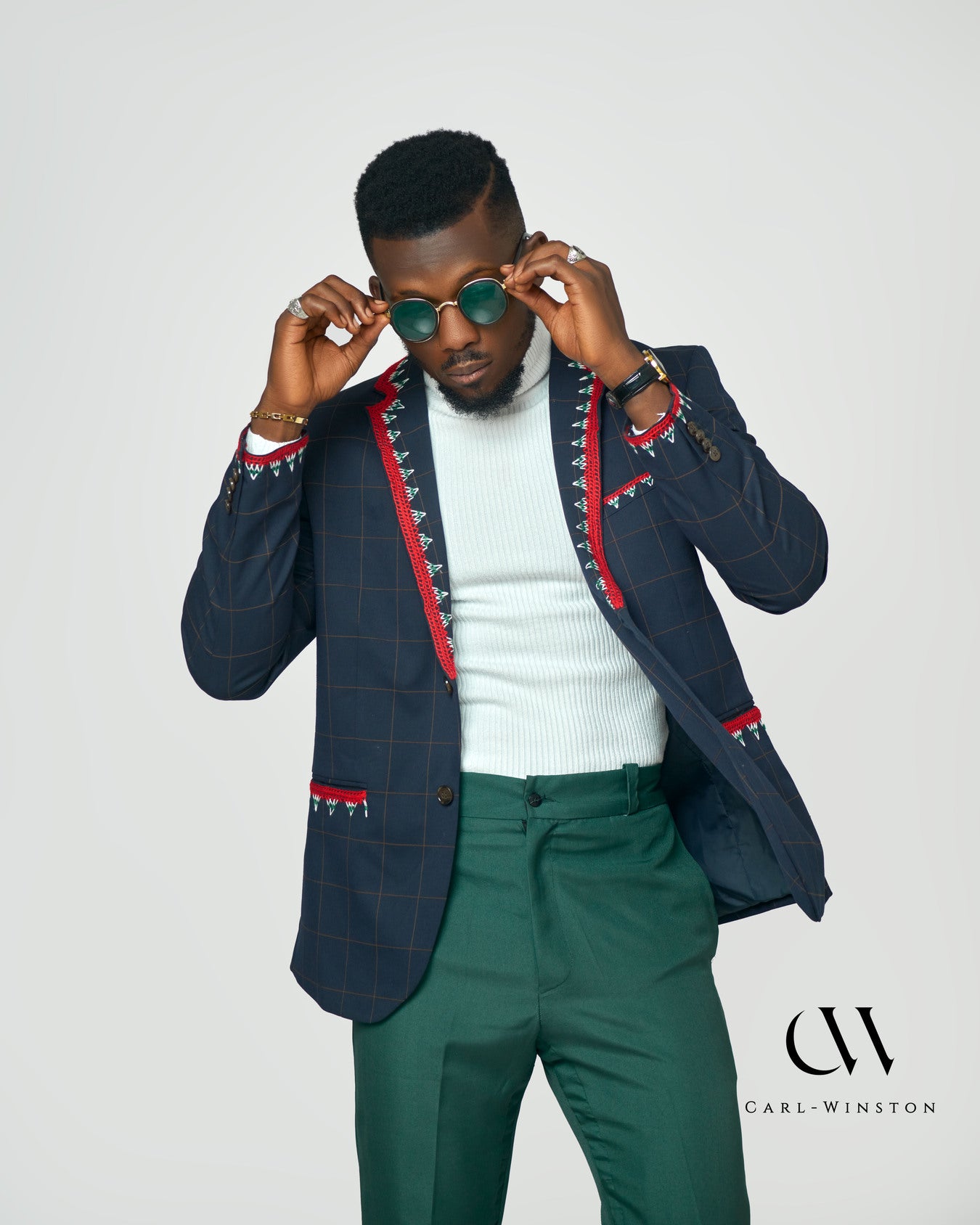 A stylish male model wearing a navy checkered blazer with intricate red and white hand embroidery, inspired by the Toghu tradition of the Tikar people. The blazer features bold cultural accents, a structured fit, and luxurious detailing.