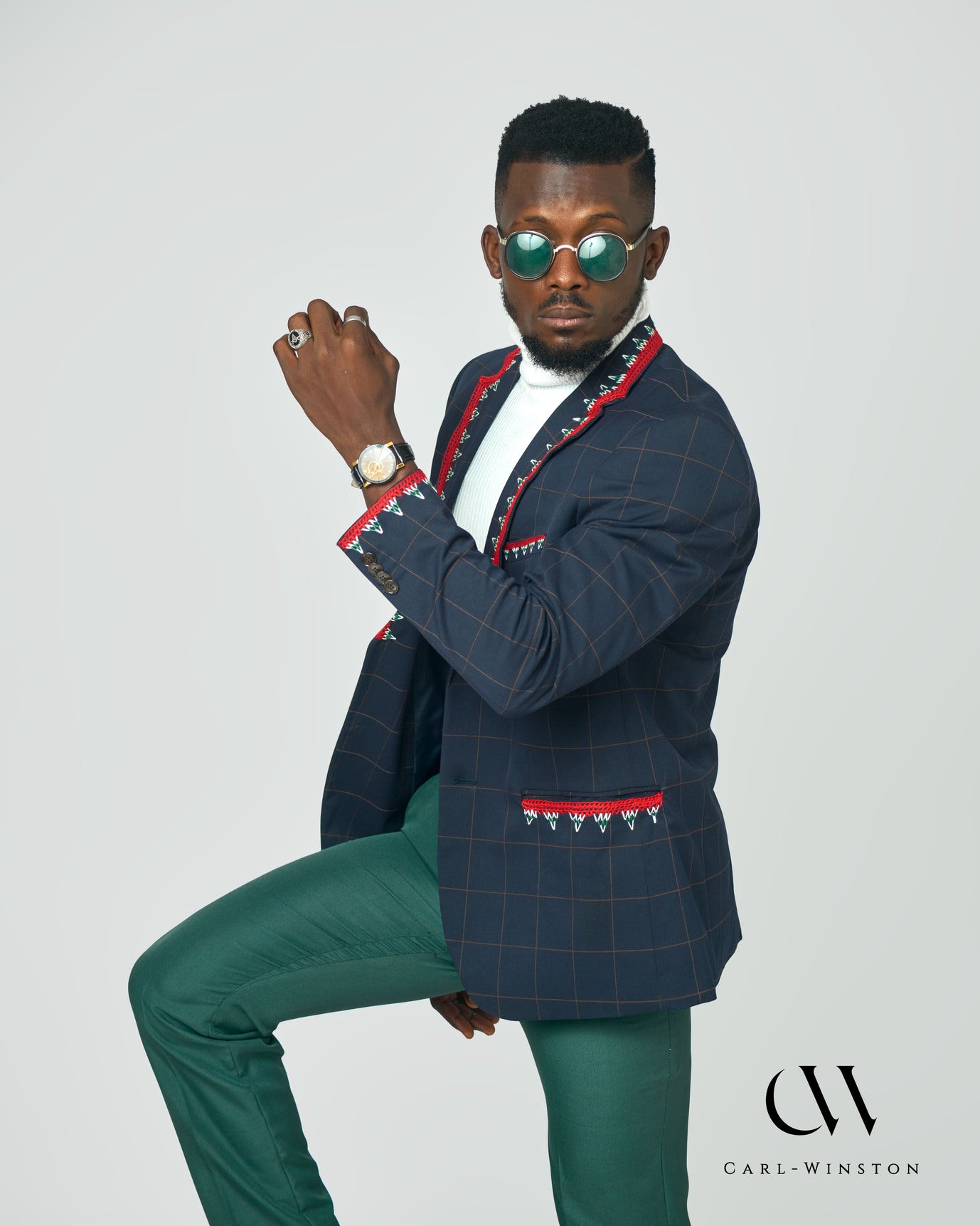 A stylish male model wearing a navy checkered blazer with intricate red and white hand embroidery, inspired by the Toghu tradition of the Tikar people. The blazer features bold cultural accents, a structured fit, and luxurious detailing.