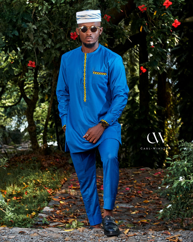 Close-up and full-body views of a male model wearing a tailored blue Elegant Atoghu Kaftan with yellow geometric embroidery, styled with matching pants and traditional hat, photographed outdoors among vibrant greenery and floral backdrops.