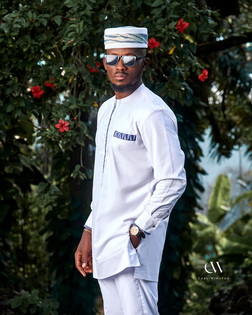 A man stands confidently outdoors wearing a pristine white Bamileke Ceremony Kaftan, designed with royal Bamileke print accents, celebrating Cameroonian heritage.