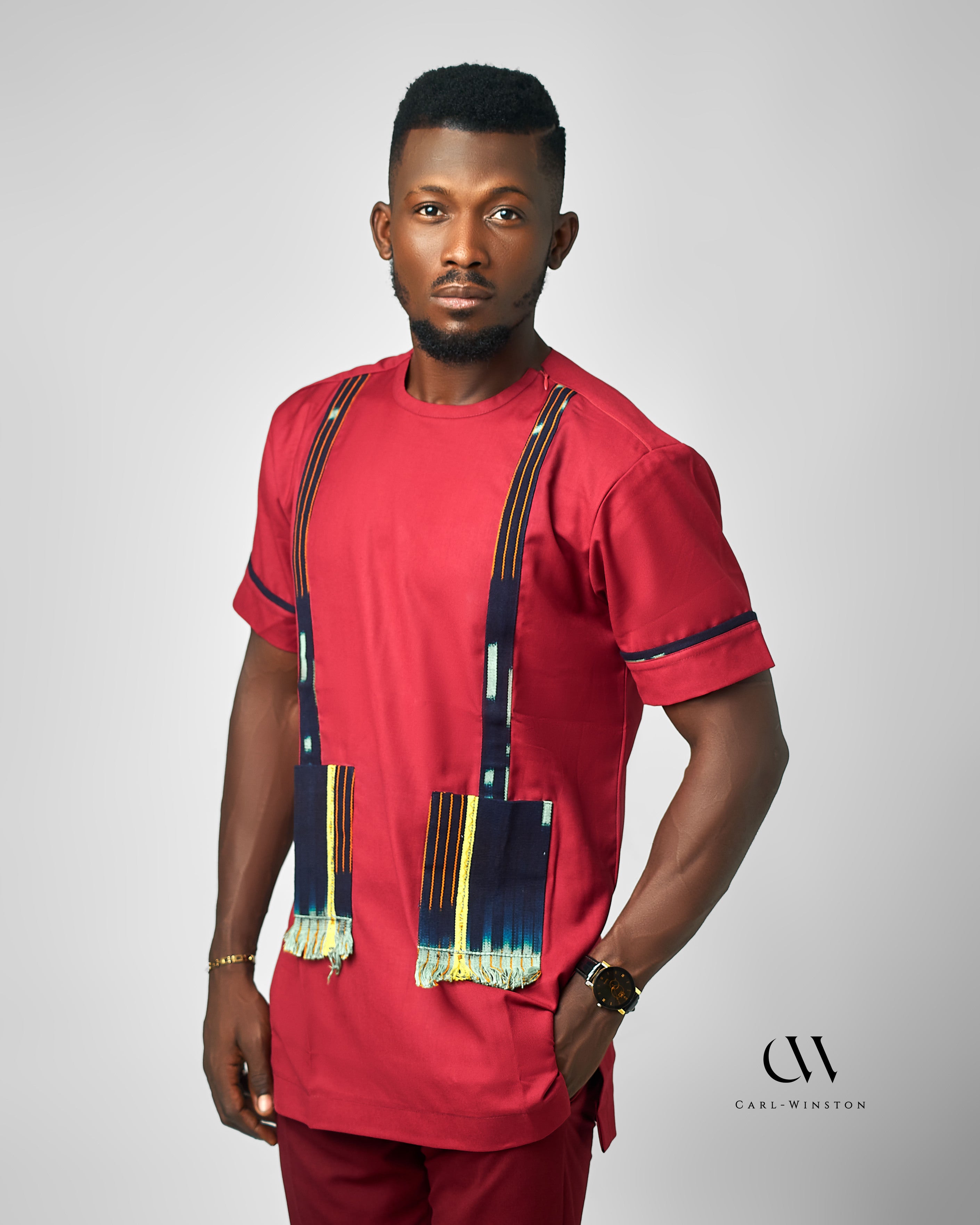 A rich red short-sleeve tunic featuring bold woven fabric pockets with fringed edges, inspired by African textile heritage. Designed by Carl-Winston, this statement piece merges tradition with contemporary style.