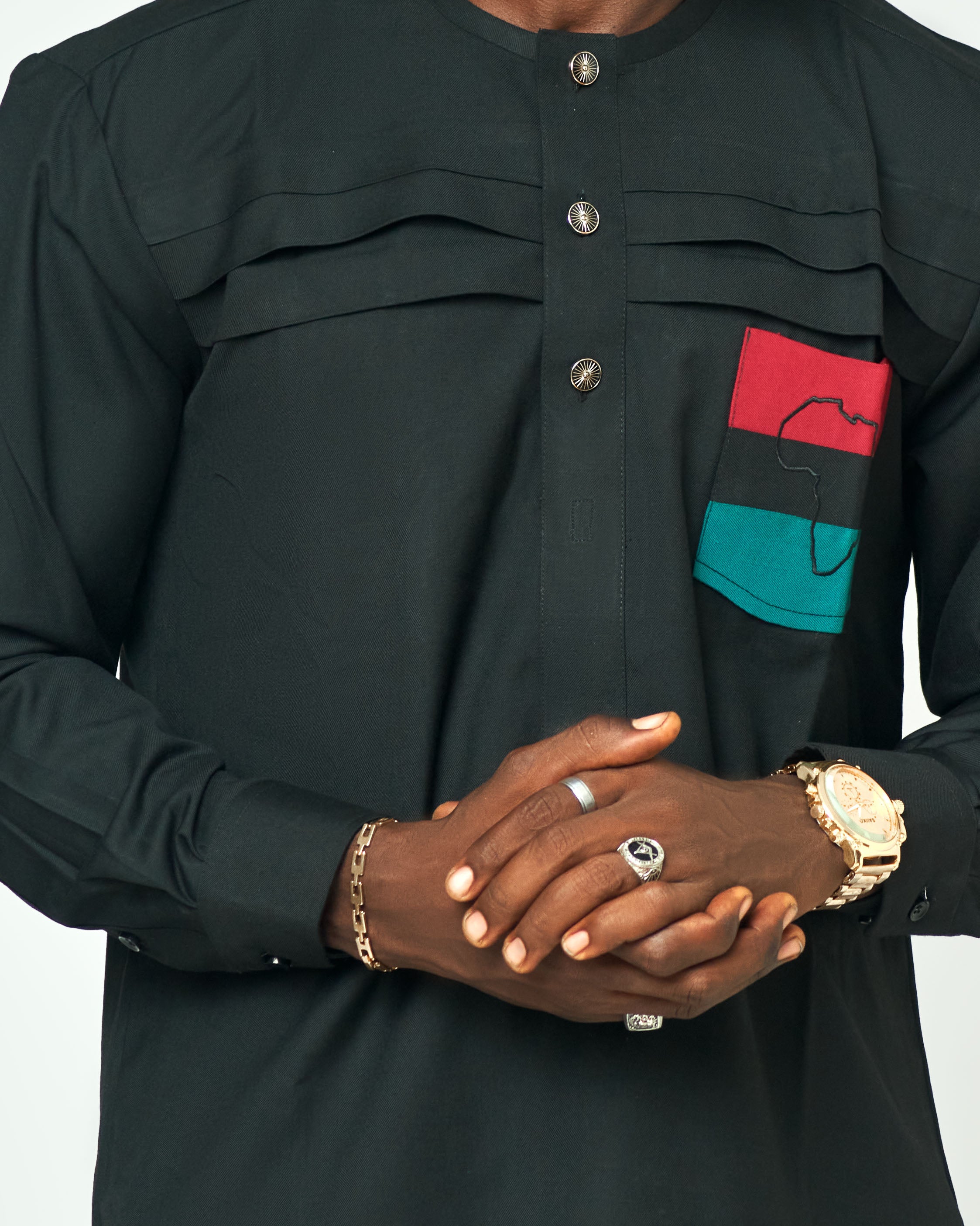 Man wearing a sleek black long-sleeve shirt with red, green, and black Pan-African-inspired accents on the pocket, paired with emerald green trousers, showcasing a modern cultural fashion statement.
