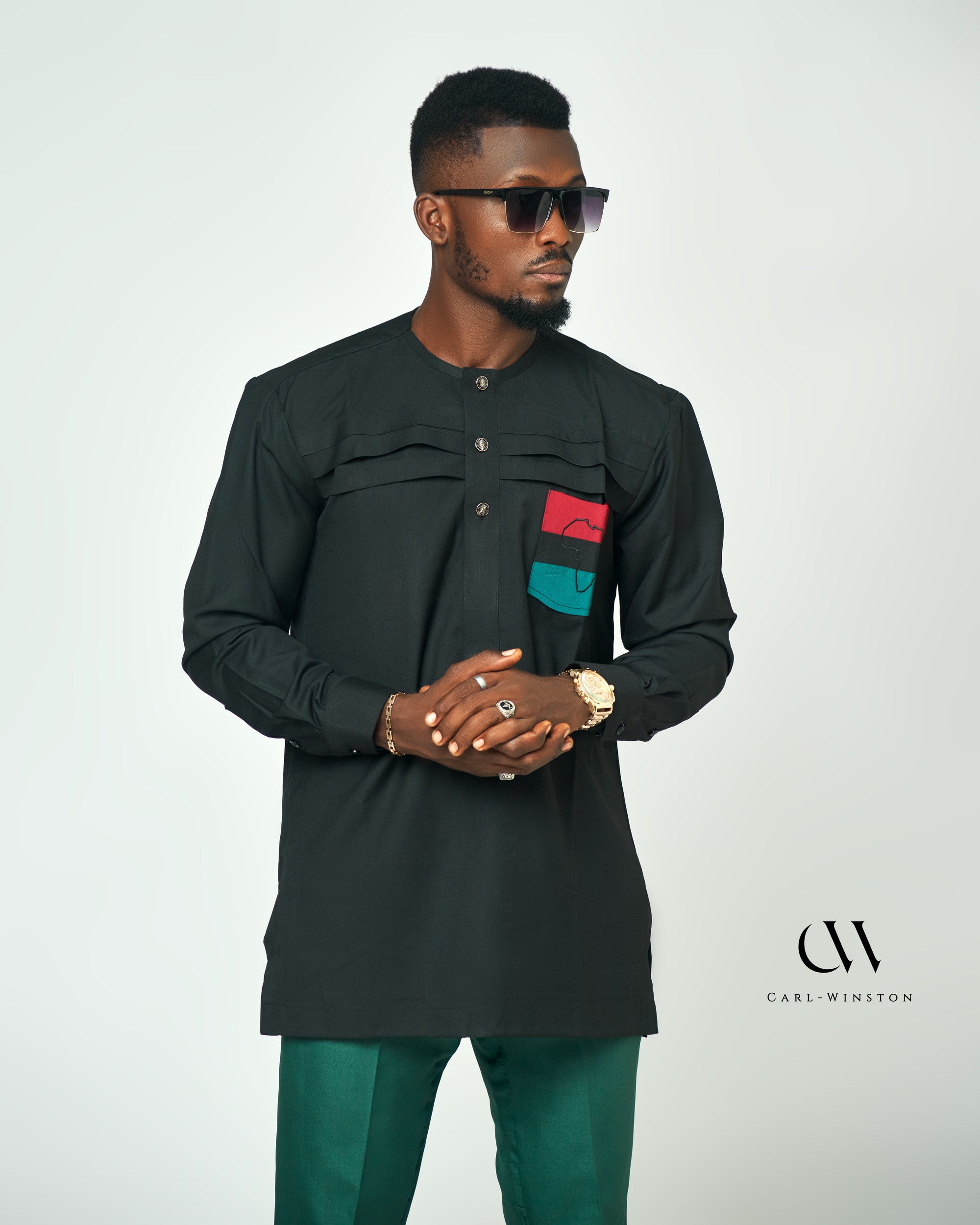 Man wearing a sleek black long-sleeve shirt with red, green, and black Pan-African-inspired accents on the pocket, paired with emerald green trousers, showcasing a modern cultural fashion statement.