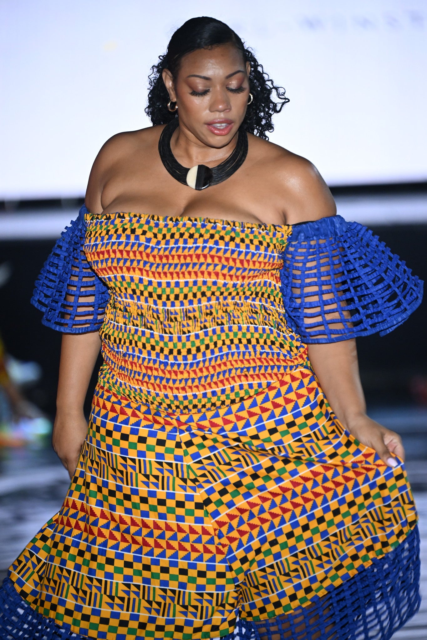 Model wearing the Carl-Winston Royal Kente Mesh Jumpsuit in XXL, featuring vibrant yellow, orange, blue, and green Ankara patterns with off-shoulder neckline, puff mesh sleeves, and mesh-trimmed hem. Styled on the runway.