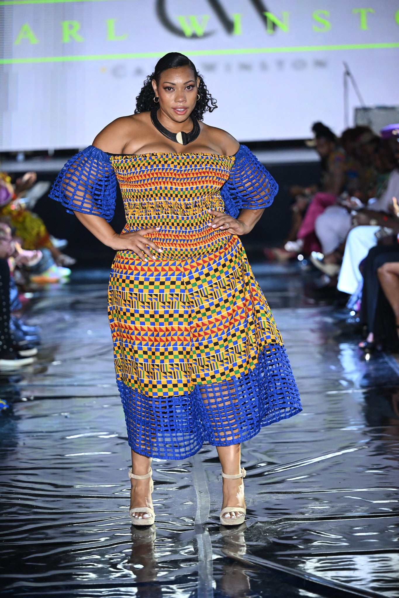 Model wearing the Carl-Winston Royal Kente Mesh Jumpsuit in XXL, featuring vibrant yellow, orange, blue, and green Ankara patterns with off-shoulder neckline, puff mesh sleeves, and mesh-trimmed hem. Styled on the runway.