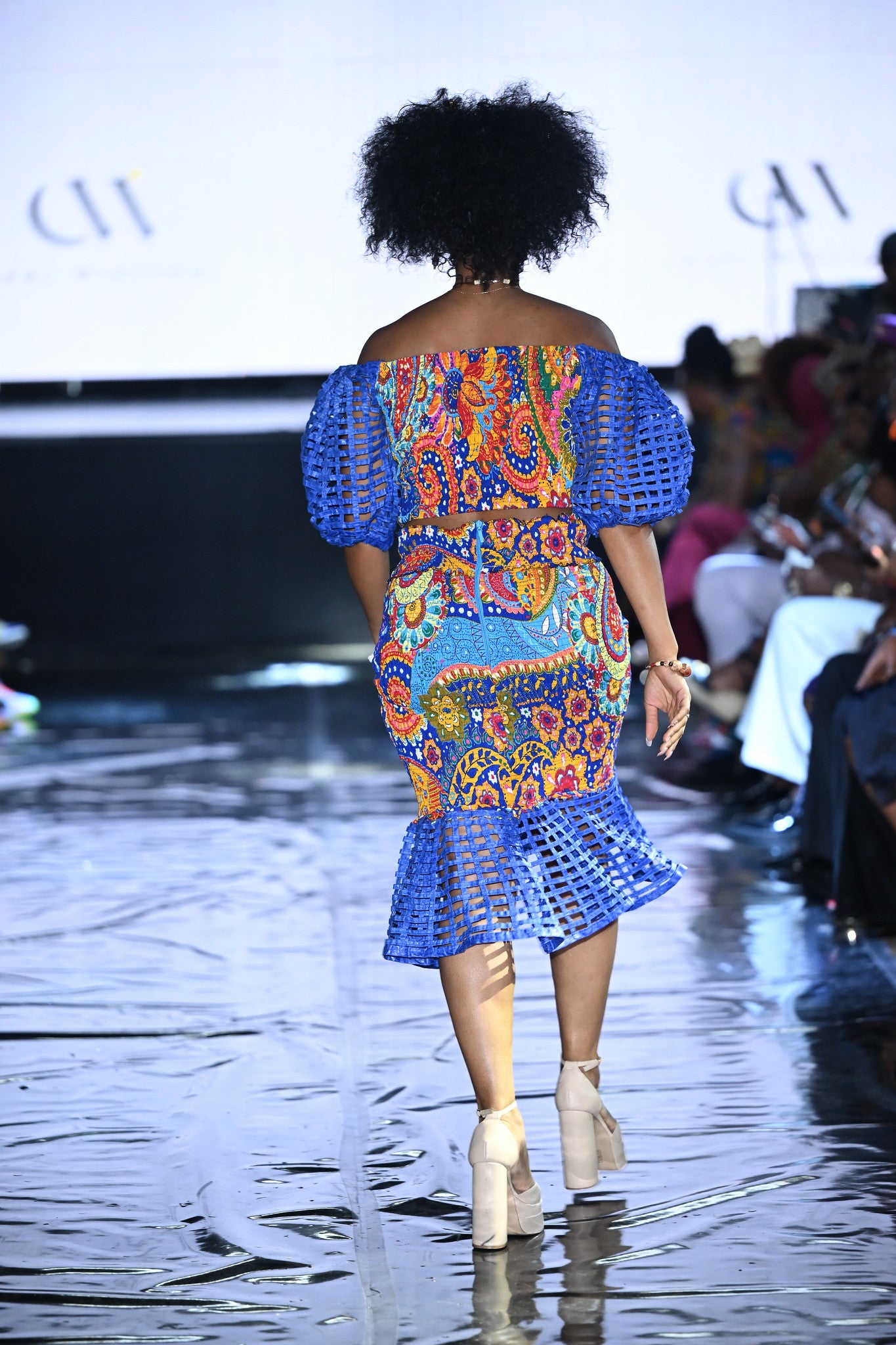Model walking on a runway wearing a colorful dress with blue sleeves and ruffled hem.