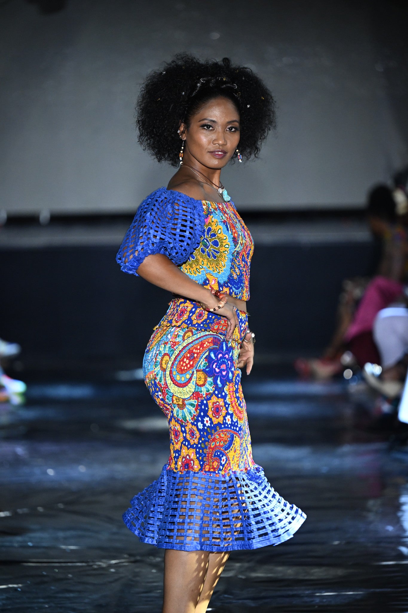 A model walks the runway wearing the Carl-Winston Royal Radiance Set: a colorful Ankara two-piece outfit featuring a fitted off-shoulder crop top with blue netted puff sleeves and a matching high-waisted skirt with flared blue hemline. The bold patterns combine orange, blue, red, and yellow in a striking modern-African fusion design.
